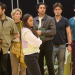 Three men and two women stand in a cluster and listen to someone off camera. They hold scripts and wear jeans shirts and jumpers.