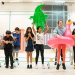 A group of children in varying heights stand in a line, a mixture of boys and girls. they are all looking distraught. One girl in the centre is holding in inflatable crocodile above her head, and another girl in the foreground is wearing a rubber ring shaped as a flamingo.