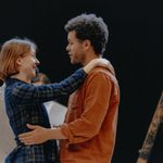 A woman and a man holding each other at arms length with happy expressions. The woman has short brown hair and is wearing a blue plaid jumpsuit. The man has brown curly hair and is wearing an orange shirt.