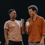 Two men holding drinking glasses and looking incredulously at each other. The man on the left has black curly hair with a fade, and is wearing a light brown t-shirt. The man on the right has brown curly hair and is wearing an orange shirt.