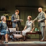 Two women and two men of different ages stand and sit on antique furniture in a living room. They all wear 1930s era clothing that is of a teal colour palette. Behind them is a piano and the wall is the same teal colour as the clothes.