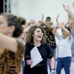 Beth Hinton-Lever with a script under their arm and a pencil behind their ear smiling in the middle of the rehearsing ensemble.