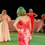 A stage in the round, grass turf of the floor with an illuminated circle. Four women are stood around, its mid wedding one woman is sat at a long banquet table looking unwell