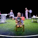 A stage in the round, grass turf of the floor with an illuminated circle. Four women are stood around all in various stages of getting ready, one kneels on the floor doing make-up