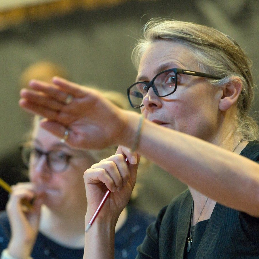 An image of Katie Mitchell in rehearsal, holding a pencil in her right hand and gesturing with her left hand towards something happening in front of her.