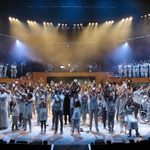 A large community ensemble onstage all lifting candles and dressed in white and cream colours