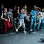 A group of people dancing during a rehearsal, being led by a facilitator in the right of the picture with a microphone