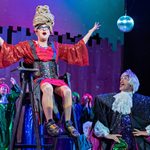 A production image from 'The Doncastrian Chalk Circle' featuring someone sat on a high chair singing and someone on the ground looking up at them, also singing.