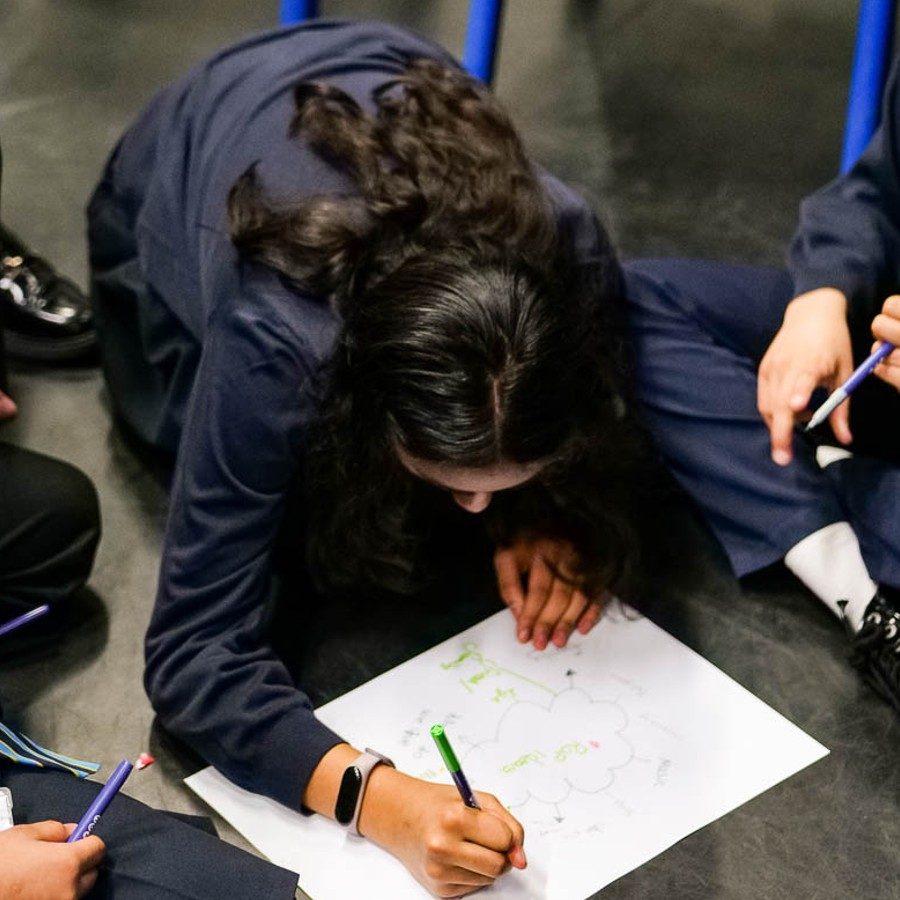 An overhead shot of a group of school students sat on the floor, one of whom is writing on a piece of paper