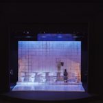 A model box showing a person sitting on a white toilet in a row of other toilets in a dirty white-tiled cubicle