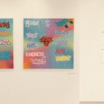 Artworks hanging on a white wall, two canvases featuring graffiti including words 'speak up', 'believe' and 'kindness'