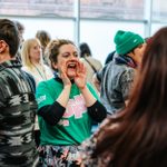 A grpup of people in a room with one person smiling and shouting in the middle wearing a green t-shirt with 'Speak Up' written on it