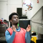 A man stands with his arms raised, his head is looking up towards a football which he is anticipating bouncing off his chest. He wears a blue long sleeved top and a red bib vest over the top.