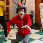 Adam Clifford, wearing a red track top with white zips, and other cast members rehearsing Hamlet for younger audiences in a room with a blue and white chequered floor.