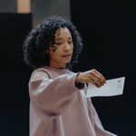 A woman with dark curly hair, wearing a pink jumper and holding a slip of paper. She is reading the paper with a disappointed expression.