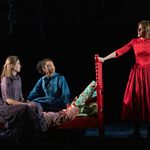 Two actresses sit on a red bed with thoughtful expressions, while a third stands confidently in a red dress, against a dark stage with hanging foliage