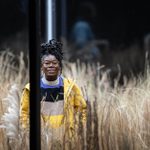 Akiya Henry, wearing glasses, a striped jumper and yellow jacket, stands amongst tall grasses