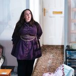 A woman is standing by a door frame, behind her is a hallway leading to a front door with a patterned carpet. In front of her is a coffee table with remotes and a box of tissues on. There is also a blue worn our arm chair with wooden handles and a pink throw pillow with ruffled edges is on it. The woman has long brown dark hair and she wears a purple dress with a gold belt chain that has sever chains hanging off it. She wears black leggings and black boots. She has red lipstick on and is looking up towards the top right.