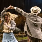 Tom Riley as Gerry spins Alison Oliver as Chris. Tom wears a suit and hat and Alison wears a blue skirt, cream blouse, and grey cardigan.