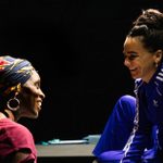 Two women, one with a colorful headdress and gold hoops, and the other in a navy Adidas tracksuit, are smiling at each other.