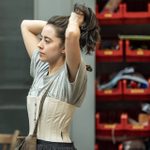 A young woman tying up her brown hair. She is wearing a corset and a brown shoulder satchel.