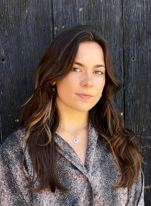 A headshot of a person with long, brown hair and wearing a patterned shirt