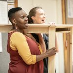 Anita Reynolds and Catrin Aaron carrying a wooden table in the rehearsal room.