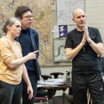 Two men and a woman watching rehearsals thoughtfully and discussing. Behind them on the wall are some large printouts and diagrams.