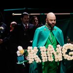 Claudius is wearing a green suit whilst holding a 'KING' funeral wreath. The rest of the cast are behind Claudius and wearing black outfits.