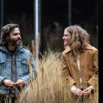 Assaad Bouab wears a denim jacket and Mackenzie Davis a light brown raincoat, both standing, looking and smiling at each other, amongst tall grasses.