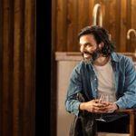 Assaad Bouab sits in a dining chair, holding a glass of wine, twisting around and smiling broadly at someone out of the picture, off to the left.