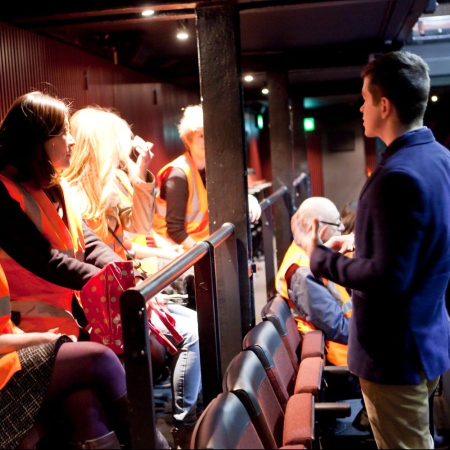 A tour guide talking to a tour group in the Dorfman theatre. Tour participants wearing orange high-visibility vests.