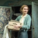 A woman in a white blouse and blue cardigan holds a wooden crate and smiles. Behind her is an antique teal coloured wardrobe that is open and lined with floral fabric.