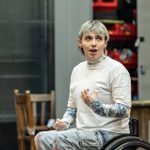 A young person with short bleached hair, wearing a white shirt over a patterned long-sleeve top, sat in a wheelchair. They are gesturing with a hand close to their chest, and singing.