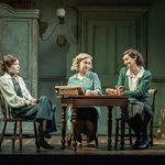 Three women in teal coloured 1930s clothing sit at a wooden table and talk. The room is all teal coloured too with a fire place and antique wardrobe.