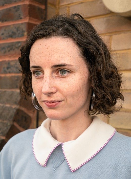 A photograph of a person with a wavy bob haircut and a wearing a white collar shit with a pale blue jumper