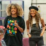 Two women stand laughing, one has shoulder length light brown hair which is very curly, the other has long brown hair that is also curly. The woman on the right wears a beret and black dungarees