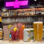 A bottle labelled Botivo, three drinks glasses with ice and slice of lime or lemon, and four cans of Small Beer and a Small Beer branded glass, filled with a light coloured beer, all lined up on a bar.