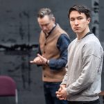 A young man with short dark hair wearing a thin grey jumper. He is frowning, with his hands clasped in front of him. Behind him is an older man in a brown waistcoat, wringing his hands.