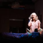 An older woman with grey hair cut into a bob sits on a bed, illuminated by a bed side lamp. She is speaking into a mobile phone that she is holding to her ear, she wears an pink night gown with short sleeves. She is holding a pamphlet. In the background you can see photos on the walls.