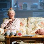 A older woman with grey hair scraped into a messy bun and secured by a orange hair scrunchie site on a white sofa with orange, yello and green floral patters. She wears a pink cardigan, a darker pink top with black leopard print spots and grey trousers underneath. In front of her is a brown wood coffee table, which has remotes, a card, coasters and a magazine. She is looking shaken whilst staring into the middle distance with her eye brows raised. She is playing absent mindly with a silver chain around her neck.