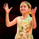 A young girl stands with her hands in the air, she is wearing a yellow dress with brown hair tired back in a bow.