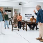 Caitlin FitzGerald, Matthew Marsh and cast members standing and sitting in a large white space, with wooden chairs and table, acting out a scene.