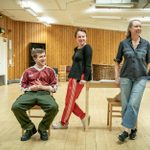 Callum Scott Howells, sitting on a wooden chair, Rosie Sheehy and Catrin Aaron leaning against a wooden table, in the rehearsal room wit hvariuos props around the room.