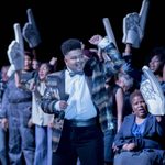 A cast member in a sparkly bowtie and suit with one fist raised in the air, holding a microphone, with the ensemble cast behind holding foam fingers