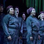 A group of cast members dressed in black boiler suits and maroon berets and a headscarf singing