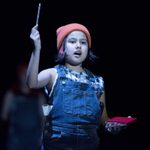 A young member of the cast in an orange hat and dungarees holding up a pencil and carrying a red notebook