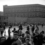 About one hundred children play in a large concrete area. There is a brutalist building in the background. Black and white photograph.