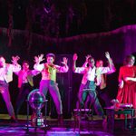 A vibrant dance scene on stage with six performers in 19th-century costume striking dynamic poses, with one actress in red observing from the side next to a table with props under a canopy of hanging foliage