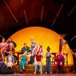 The company sit across the stage singing and playing instruments. Behind them is a large yellow sunset lighting up the stage.